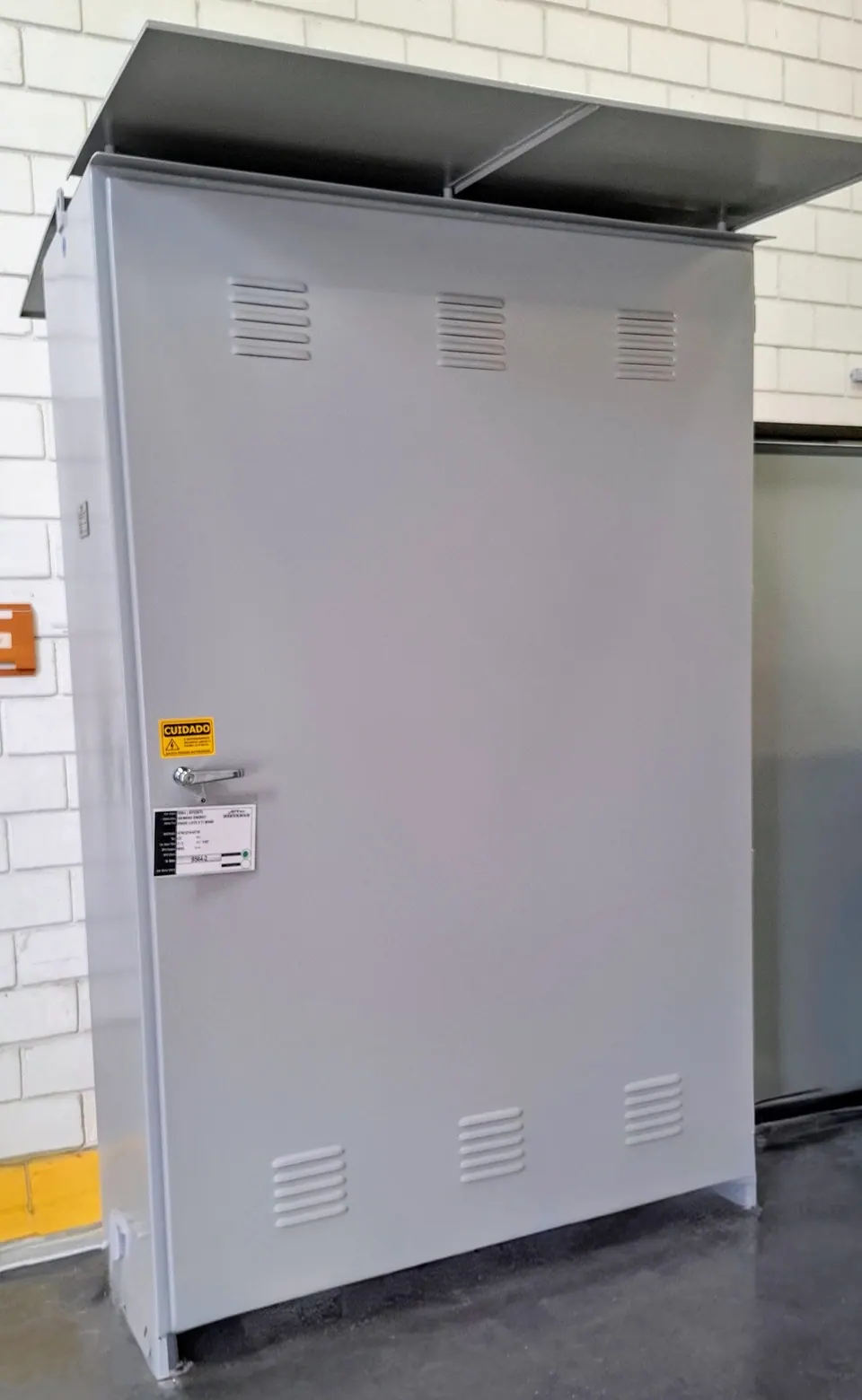 Armário de transferência de reator reserva fechado, em chapa cinza-clara, com cobertura no teto, venezianas na porta e placa amarela de advertência
