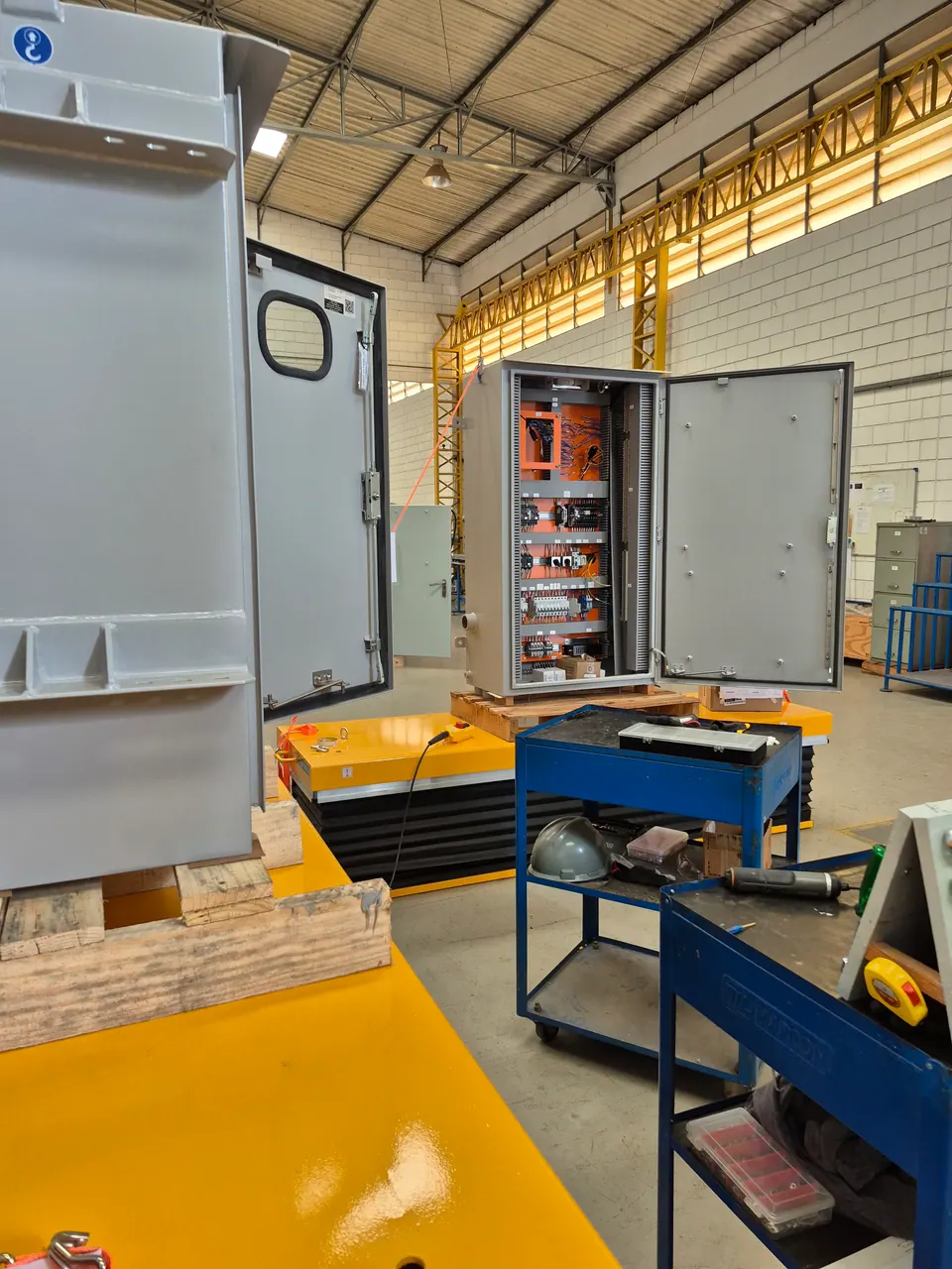 Painel de controle aberto sobre uma mesa elevatória no galpão de montagem, com a placa laranja já fiada; em primeiro plano, outro gabinete com a porta aberta