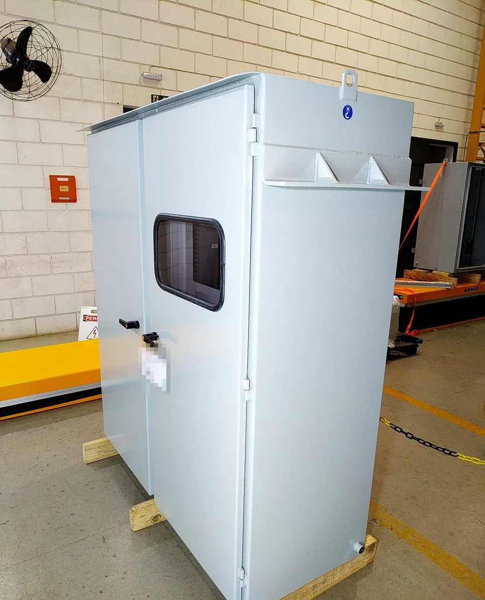 Closed light-gray cabinet on a wooden skid on the factory floor, with two doors, a rectangular viewing window in the right door, black handles, and a lifting eye on the roof